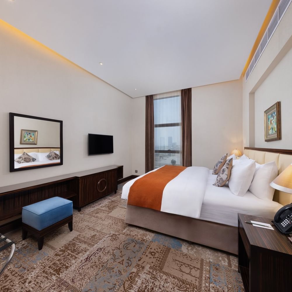 Orange bed throw by a blue velvet ottoman on a patterned rug under a wall-mounted TV in Executive Room at Warwick Al Khobar