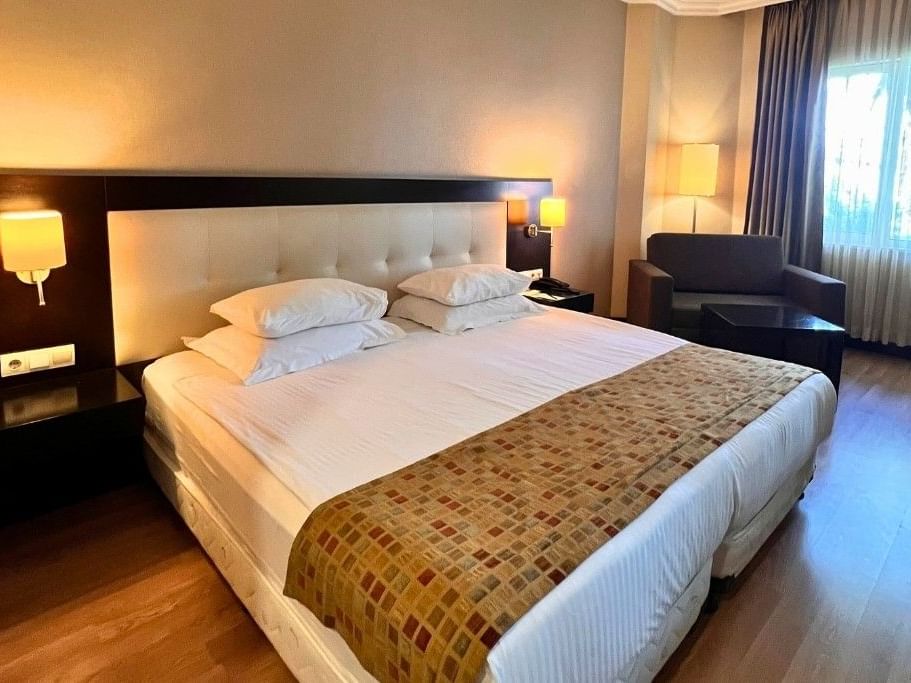 A hotel room with a large bed, four pillows, nightstands, lamps, armchair, and a window with curtains.