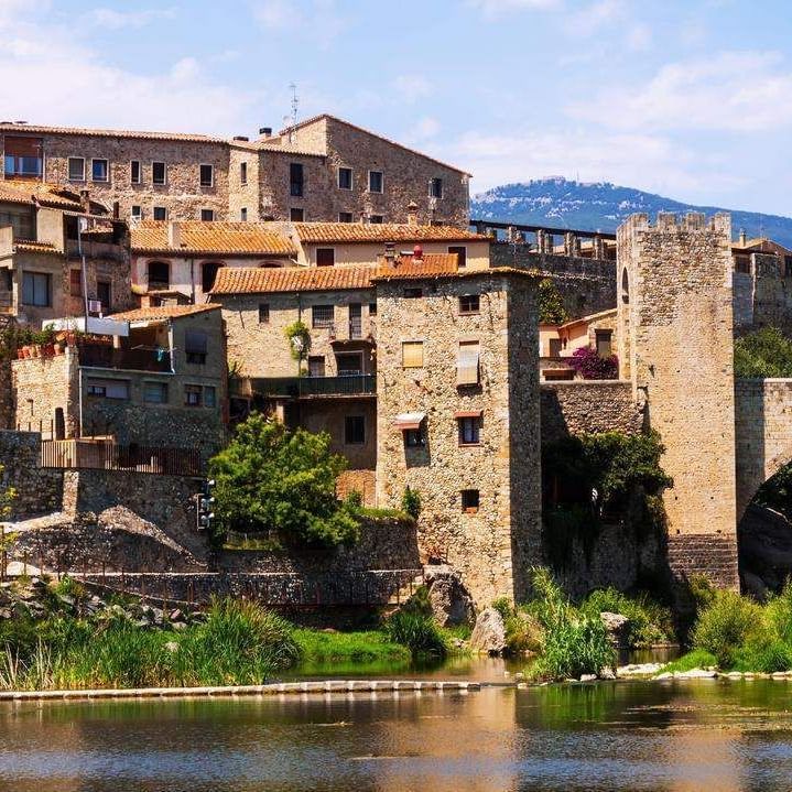 Stone bridge and medieval buildings beside a river with mountains in the background.