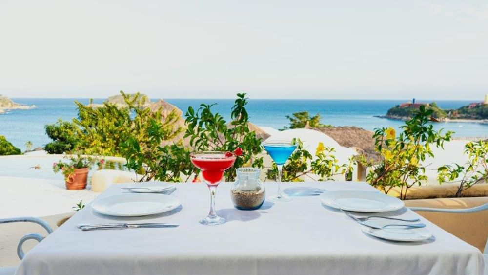 Cocktails on a white cloth table by green plants under a clear sky with a view at Quinta Real Huatulco