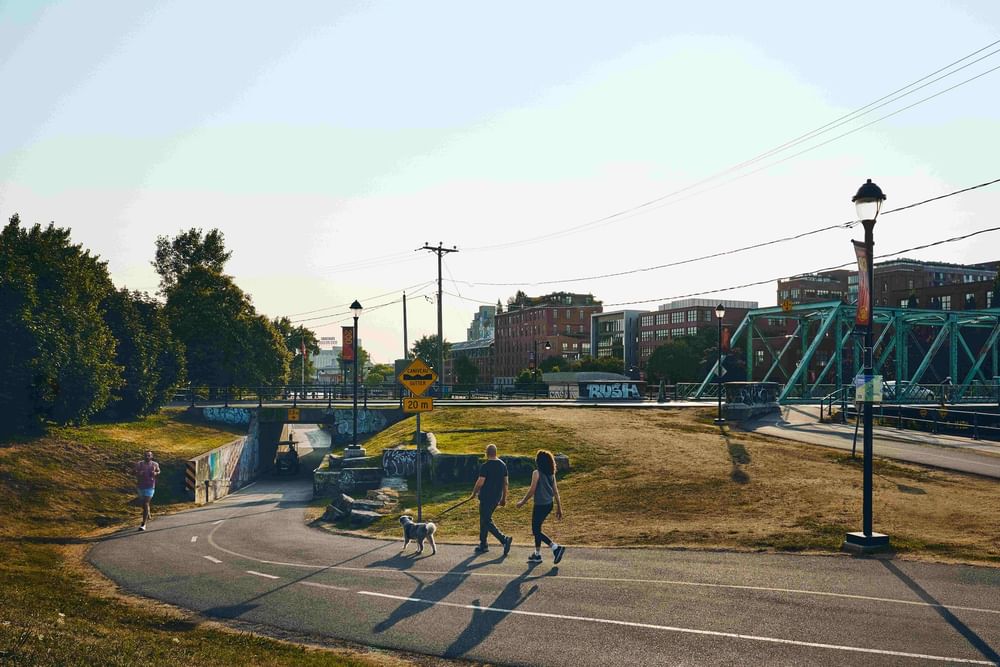 People walking with a dog on a paved path near a green bridge and urban buildings at Warwick Le Crystal Montreal