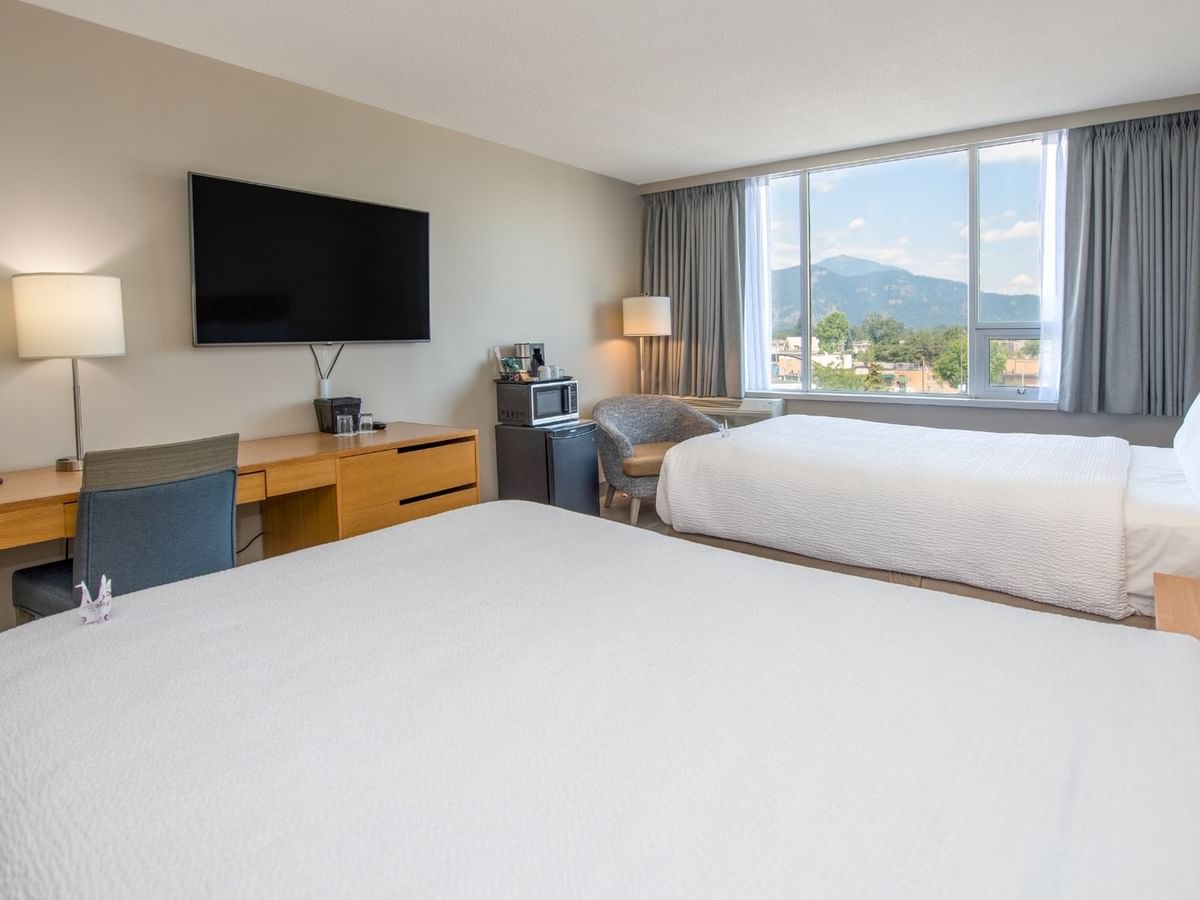 Two beds, nightstands, TV, and work desk near a window with a mountain view.
