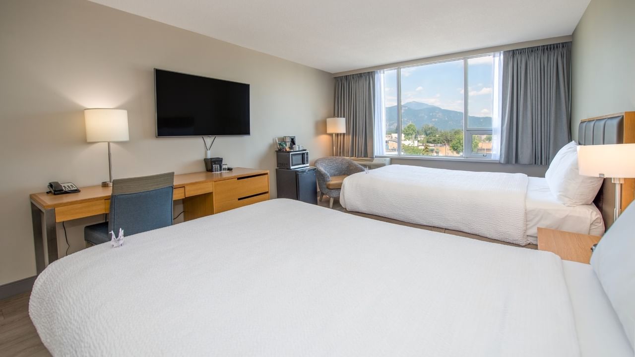 Two beds, nightstands, TV, and work desk near a window with a mountain view.
