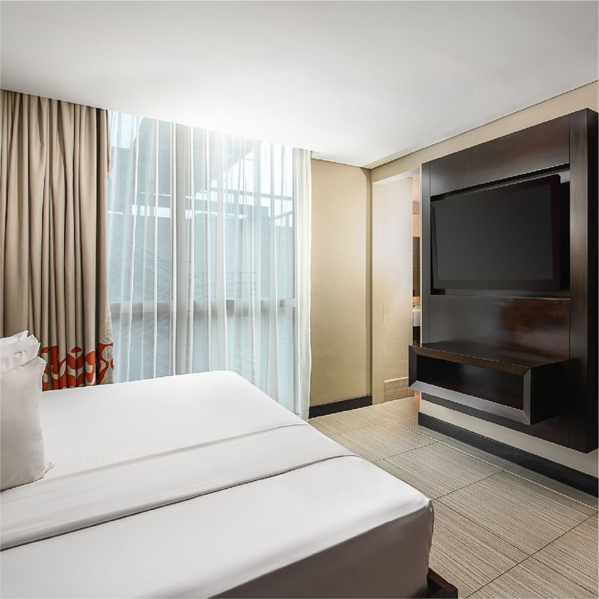 Dormitorio moderno con cama blanca y ventana con visillos frente a una pared con televisor en Megapolis Hotel Panama