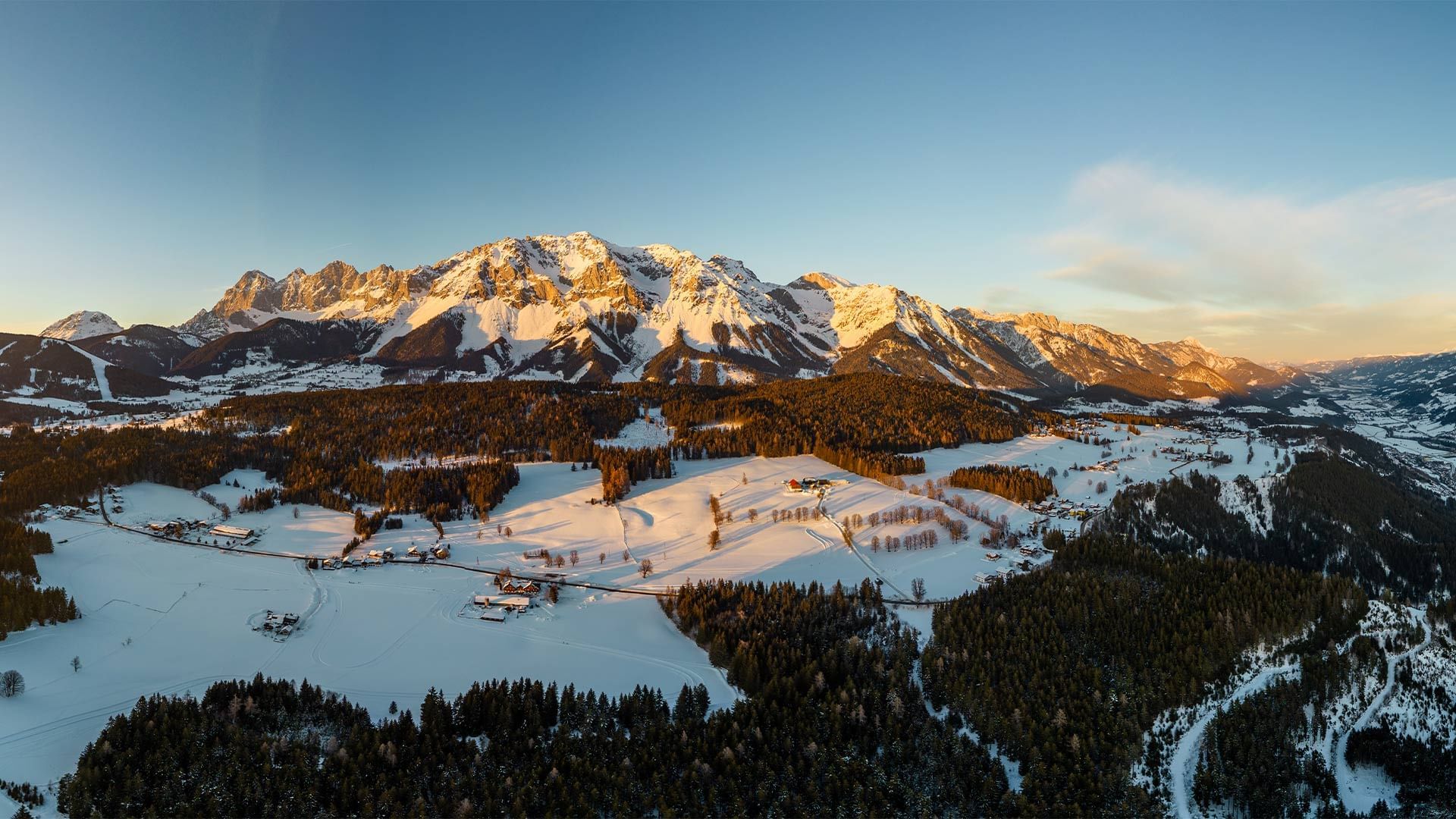Aerial view of snowy valley and mountain range with clear sky and a few scattered clouds.