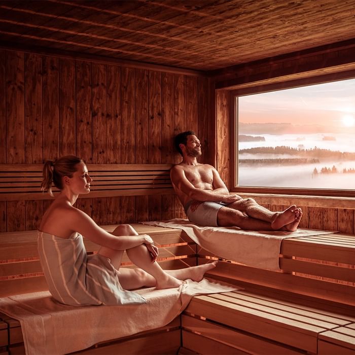 Coppia rilassata in una sauna di legno con vista sulla foresta e nebbia mattutina.