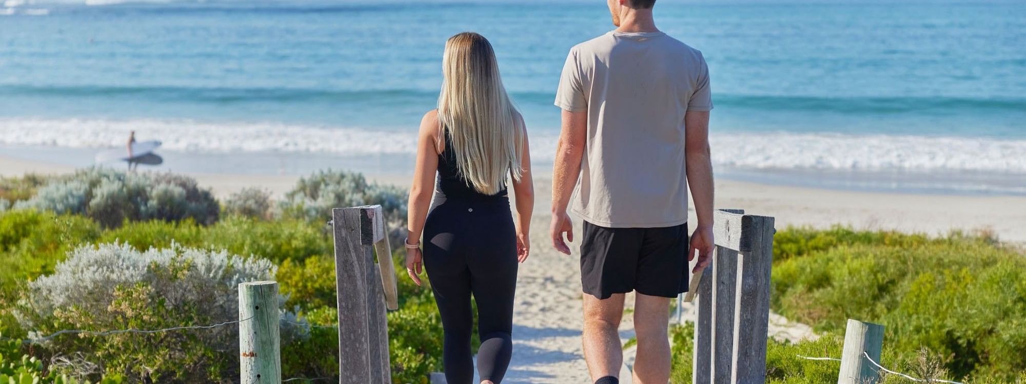 Couple walking on wooden boardwalk near beach with ocean view, promoting Stay Longer, Save More offer.