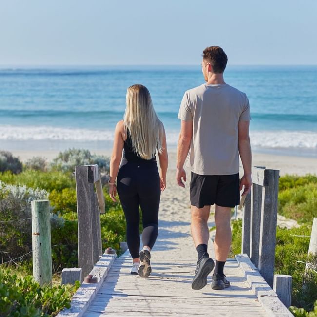 Couple walking on wooden boardwalk near beach with ocean view, promoting Stay Longer, Save More offer.