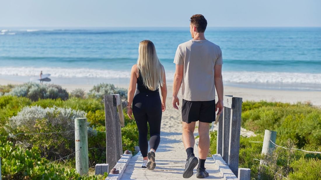 Couple walking on wooden boardwalk near beach with ocean view, promoting Stay Longer, Save More offer.