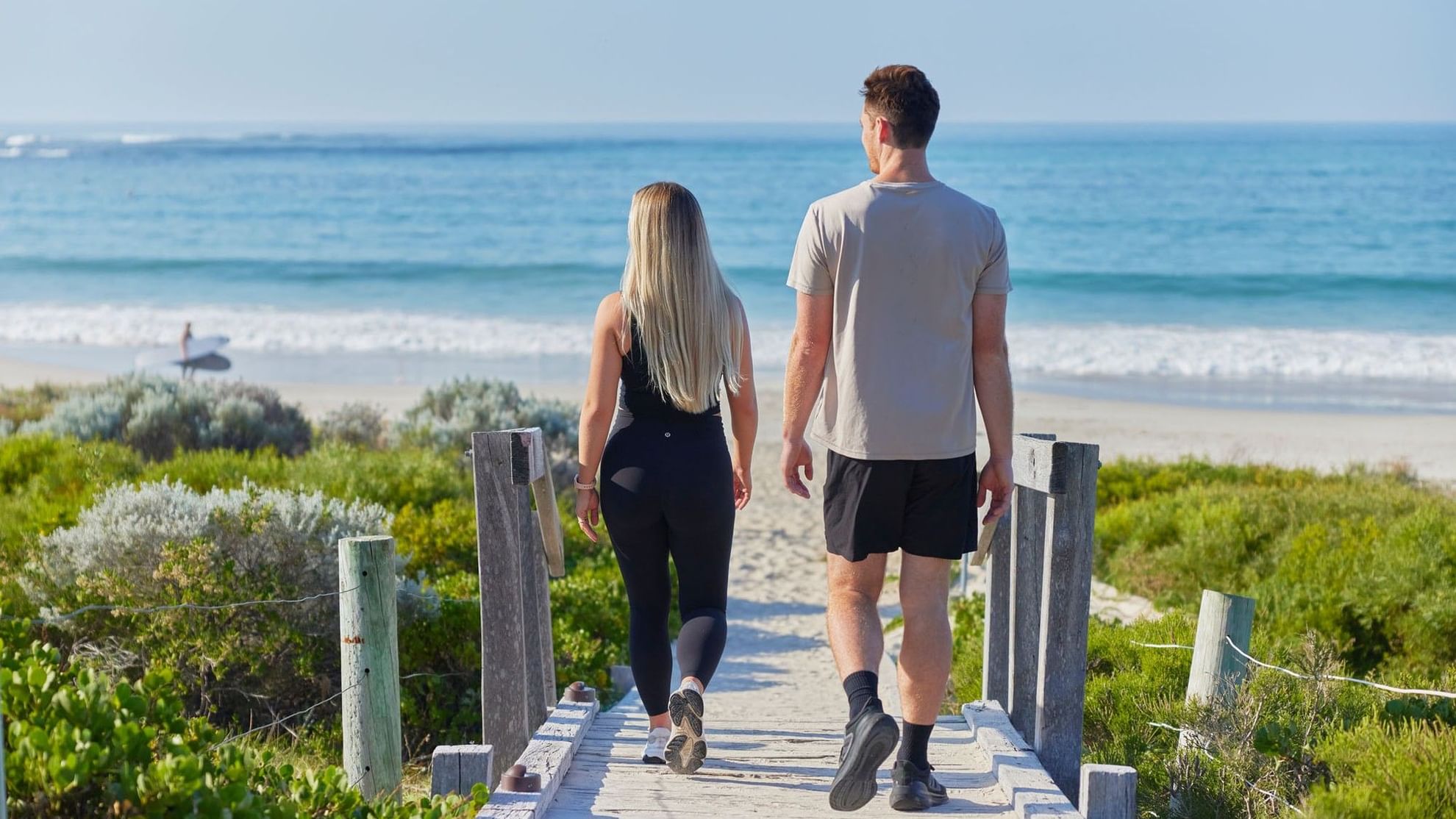 Couple walking on wooden boardwalk near beach with ocean view, promoting Stay Longer, Save More offer.