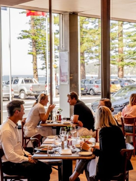 Breakfast at Elysium Manly with diners by tables under large windows surrounding a view of pine trees and street traffic