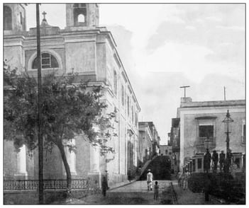 Black and white image of a street with several people walking near a church and other buildings.