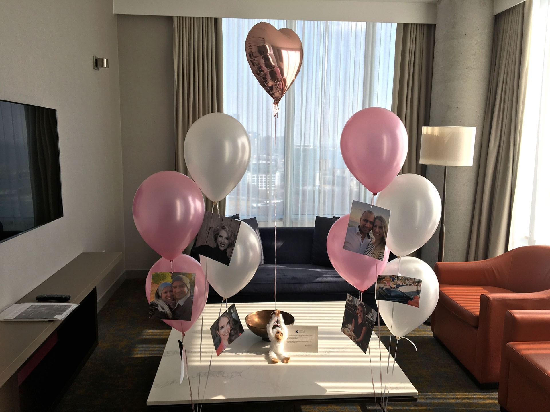 Pink and white balloons with photo cards floating by a navy sofa and window, near a rose gold heart at Hotel X Toronto