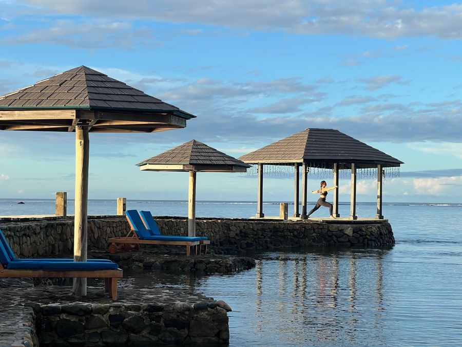 Woman practicing yoga on a platform by the ocean with gazebos and lounge chairs at Warwick Fiji Resort and Spa.