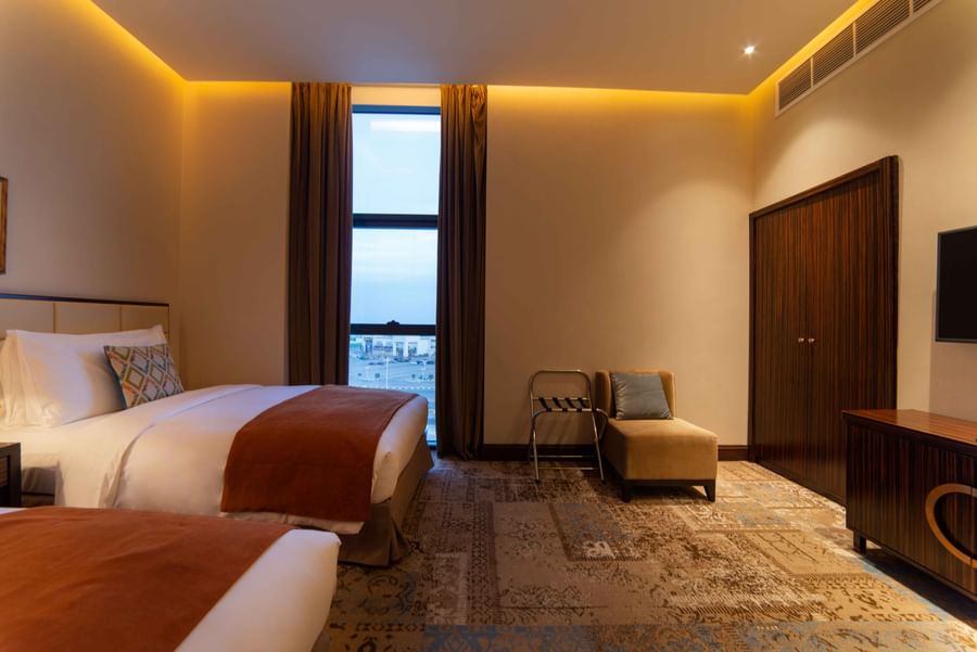 Two Bed & Single armchair with a gray pillow by a tall window,dark wood wardrobe on a patterned rug at Warwick Al Khobar