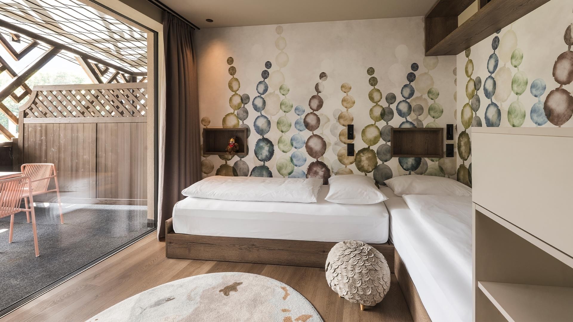 A cozy bedroom with patterned walls, two beds, a round rug, and a glass door leading to an outdoor area.