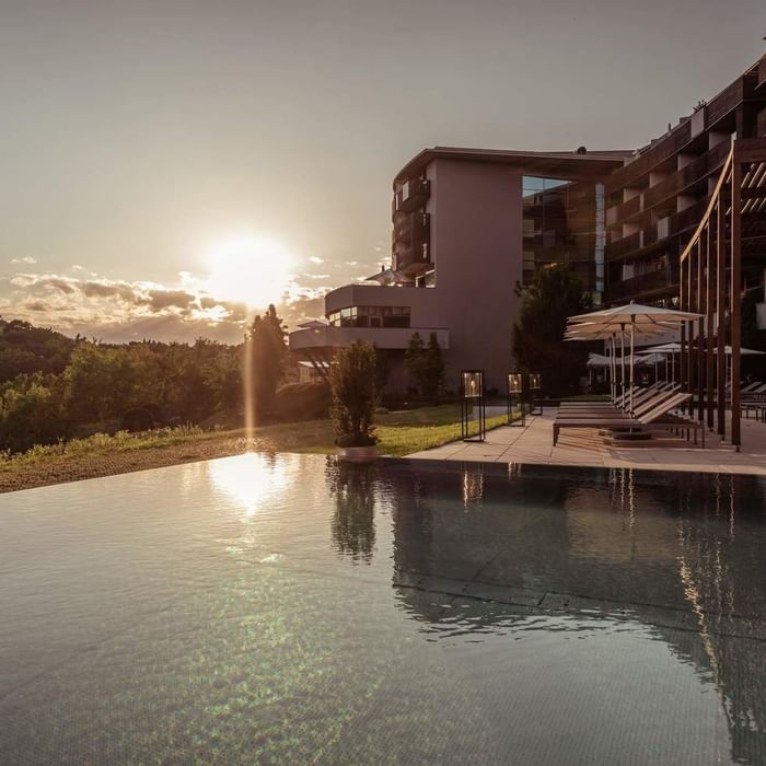 Poolbereich mit Liegen und Sonnenschirmen vor einem Hotelgebäude bei Sonnenuntergang.