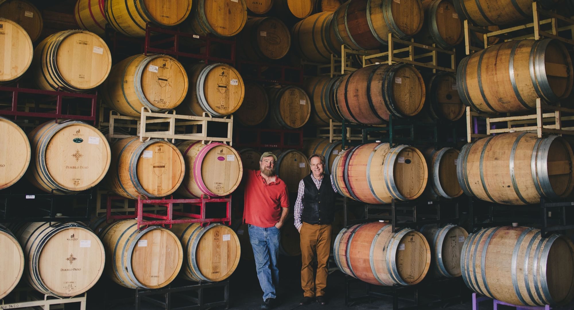 Two men standing by stacks of wine barrels 