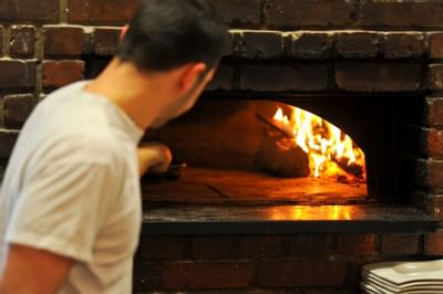 Man tending to a blazing fire inside a brick pizza oven in Piccolo Arancio at Farmington Inn and Suites