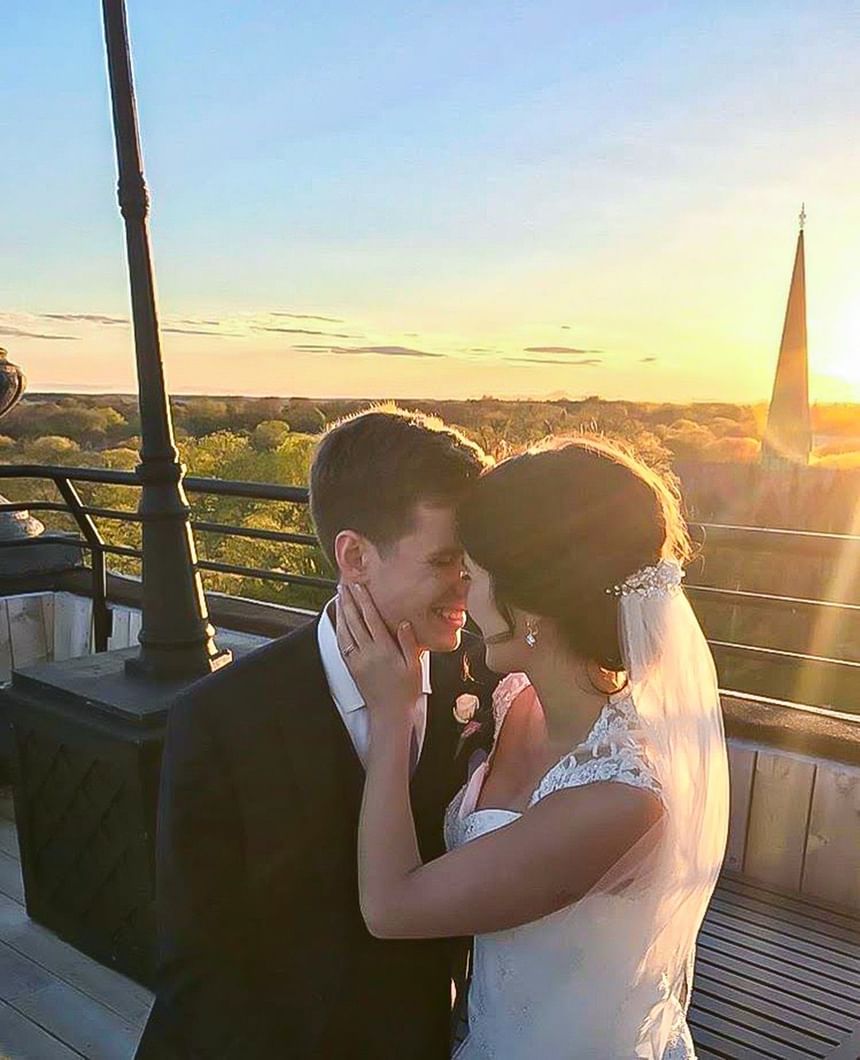 Bride and groom smiling by a balcony railing under a warm sunset with a church spire at Rodd Charlottetown Hotel