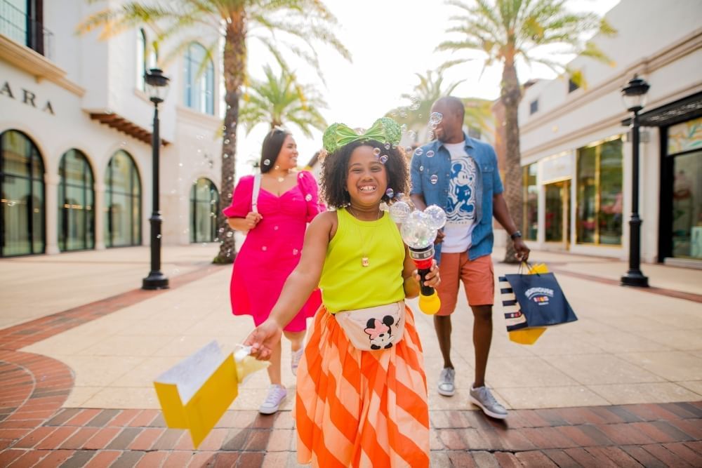 A girl in a yellow shirt and orange striped skirt walks in front of two adults swinging shopping bags between storefronts and palm trees. 