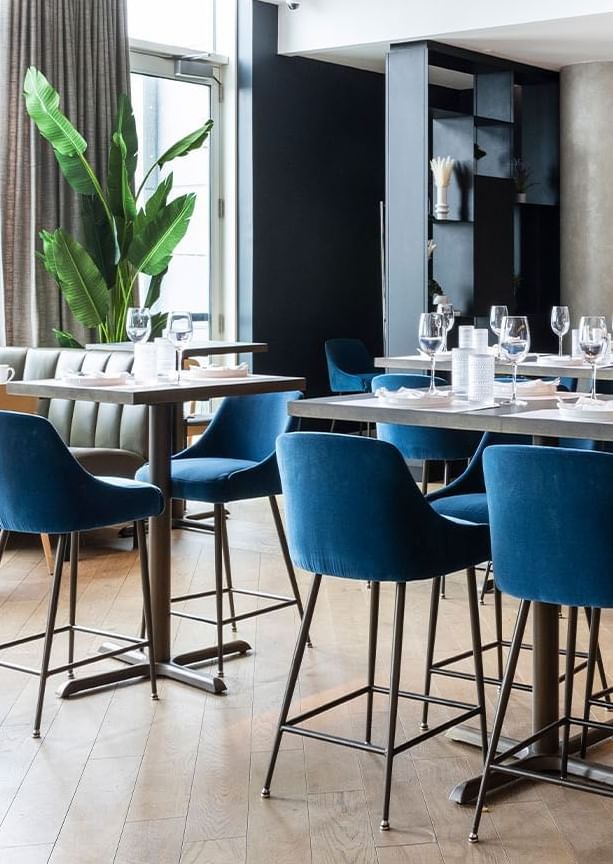 Roses Social dining area with tall bar stools facing wooden tables beside tall green plants and shelves at Hotel X Toronto