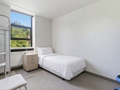 White room with single bed, chair, and window view of trees at UniLodge Stafford House.