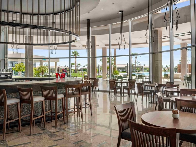 Bar and dining area with round tables, chairs, and large glass walls showing outdoor patio and pool.
