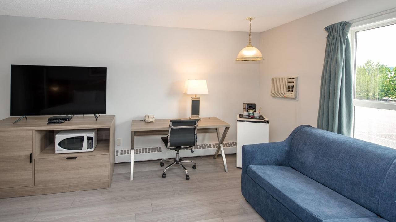 Room with blue couch, desk with chair, TV, microwave, lamp, and window with curtain.