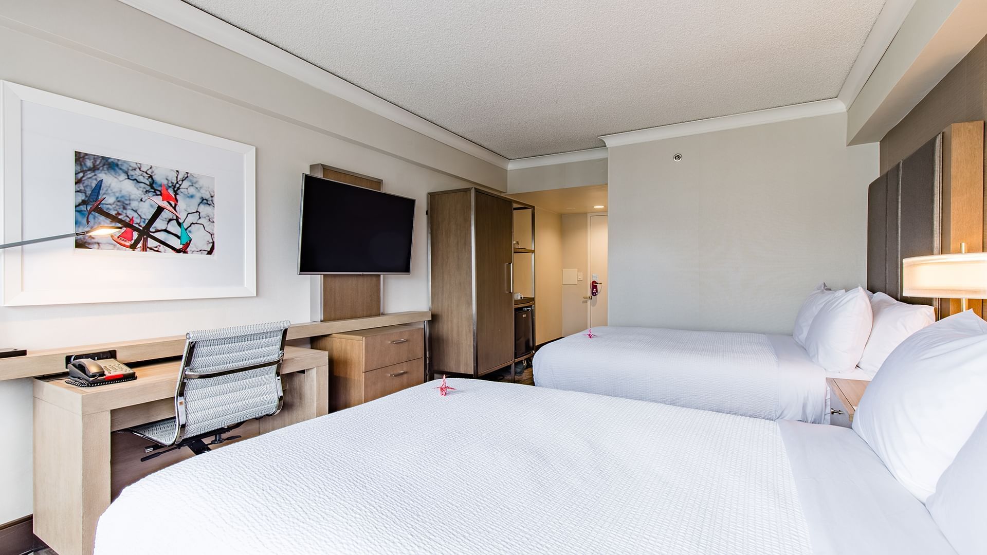 A hotel room with two beds, a desk, TV, and artwork.