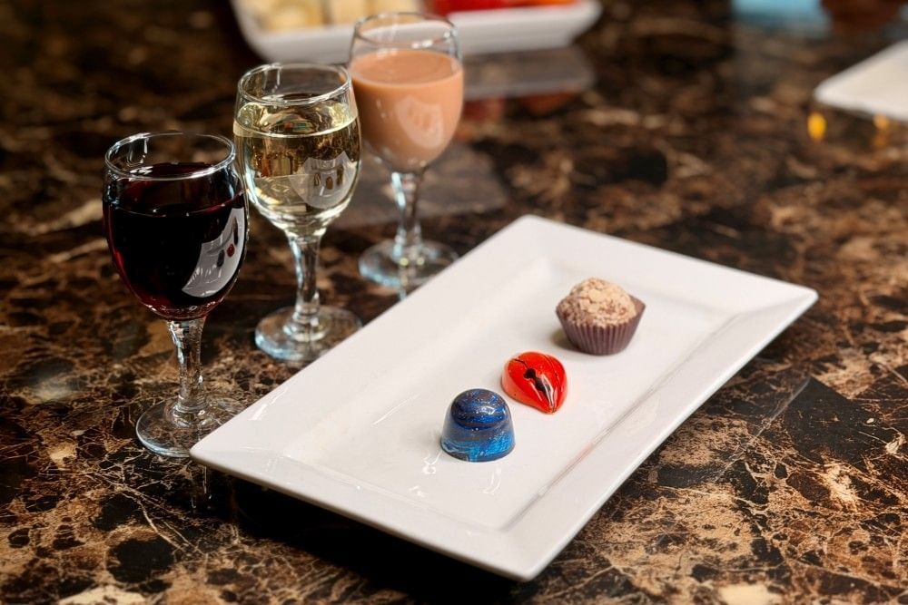 Three glasses of wine sit on a dark granite table beside a rectangular white plate adorned with three colorful chocolate truffles.