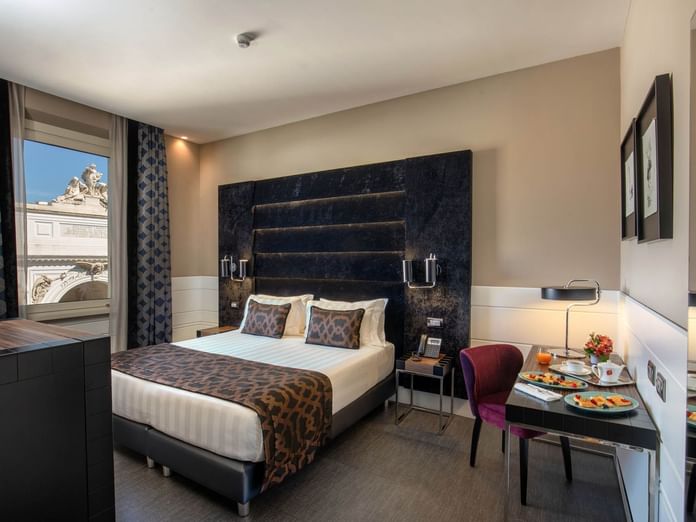Bed beside a desk with a breakfast setup and large windows offering a scenic view in the Superior Room at The Glam Hotel