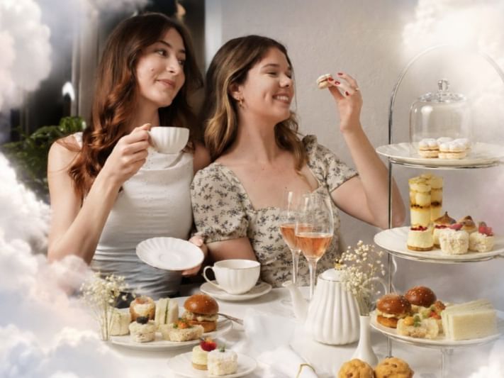Two women enjoying Cloud High Tea with tiered pastries, sandwiches, and tea in an elegant cloud-themed setting.