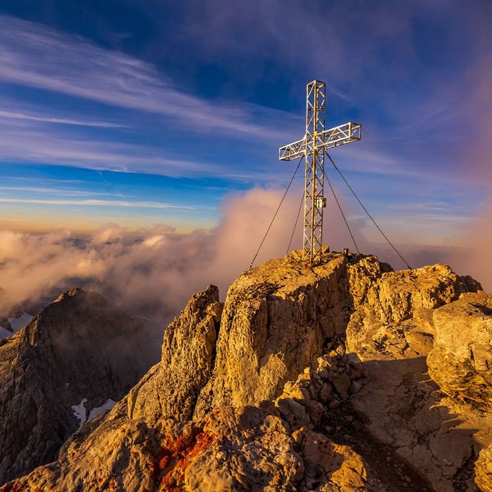 Croce di metallo su vetta rocciosa con cielo blu e nuvole rosa al tramonto su Falkensteiner Hotel Schladming