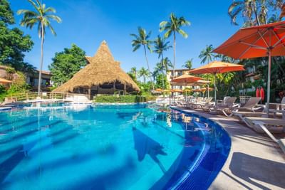 Blue pool with lounge chairs and umbrellas, hut, and palm trees on a sunny day.
