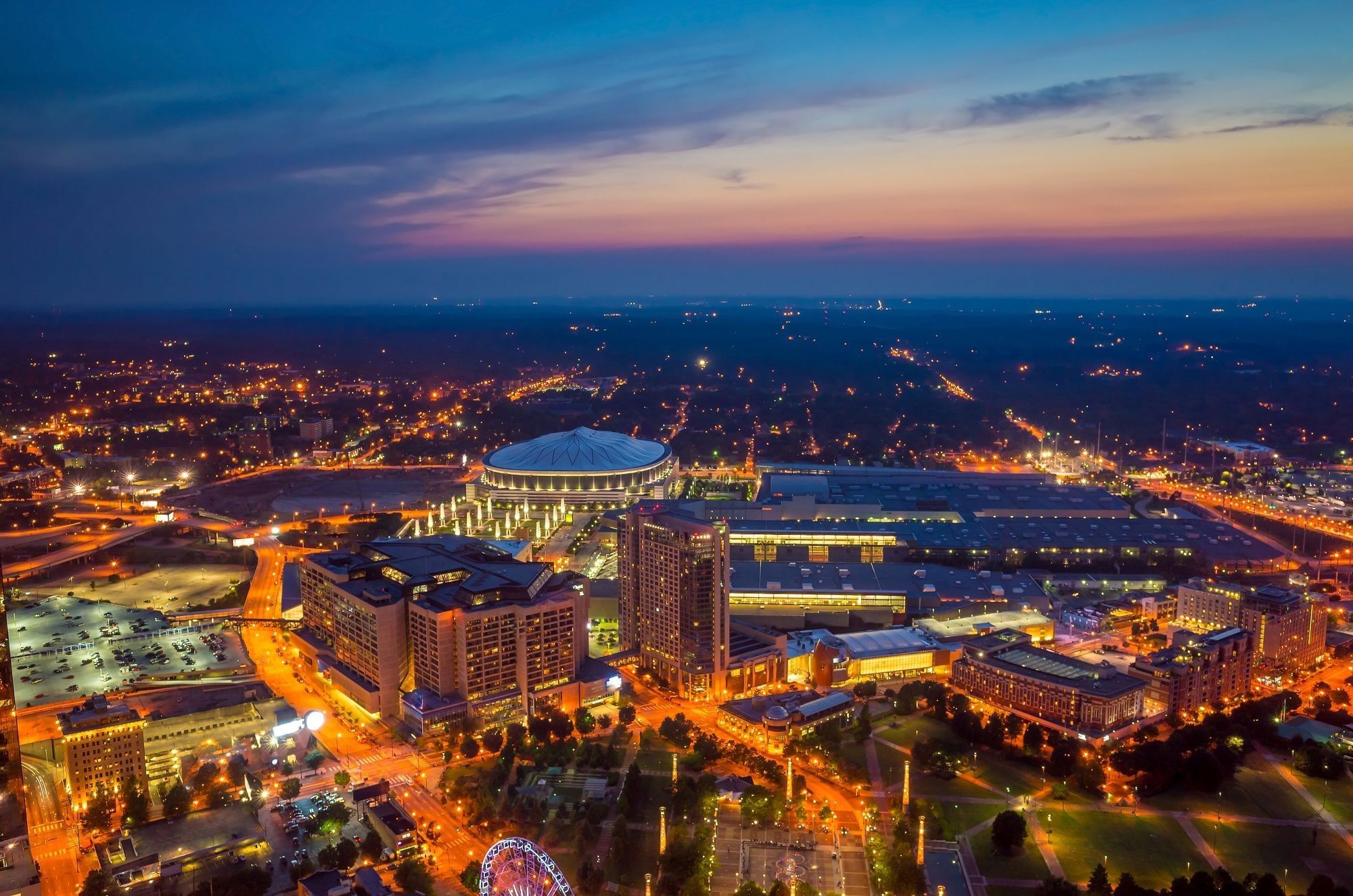 An aerial view of a city at dusk with a large building complex and a dome-shaped structure.