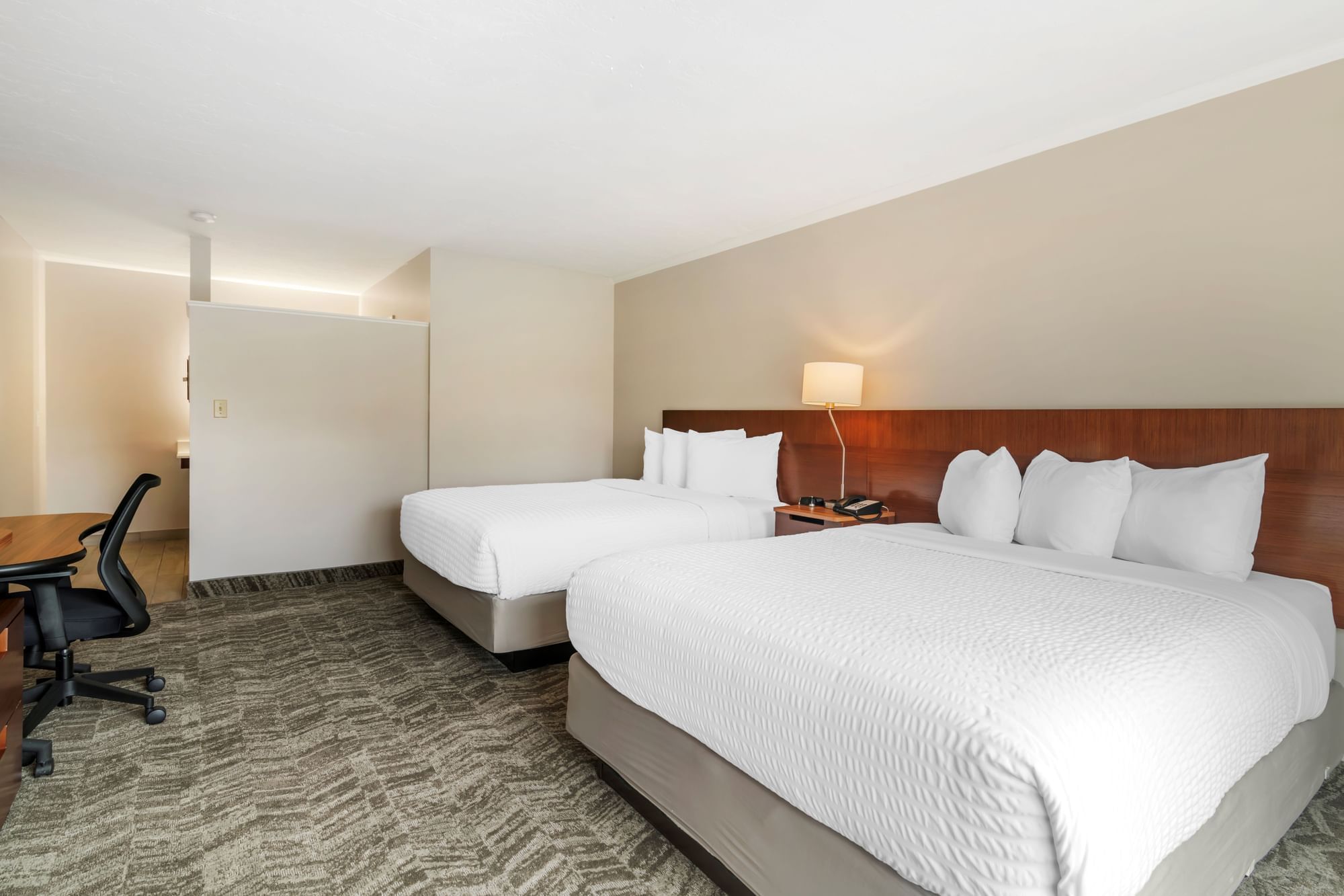 A hotel room with two queen beds, a desk, and a lamp.