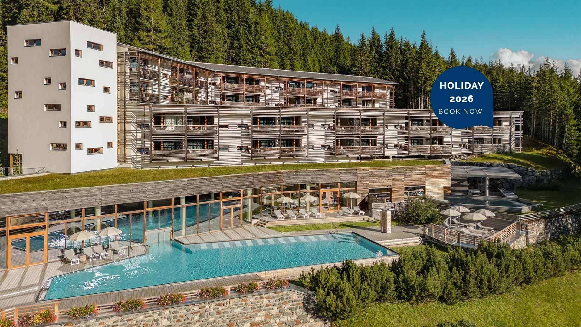 Modernes Hotel mit Pool und Liegen, umgeben von Wald und Wiesen, mit Werbetext HOLIDAY 2026 BOOK NOW!