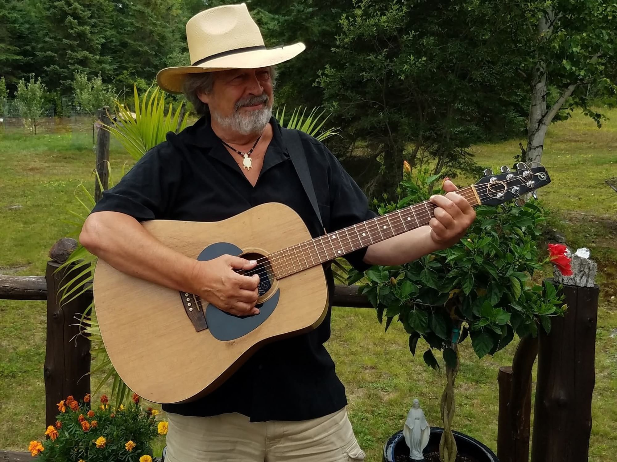 Wayne Failing in a cowboy hat playing a guitar by a wooden fence, surrounded by green garden plants.