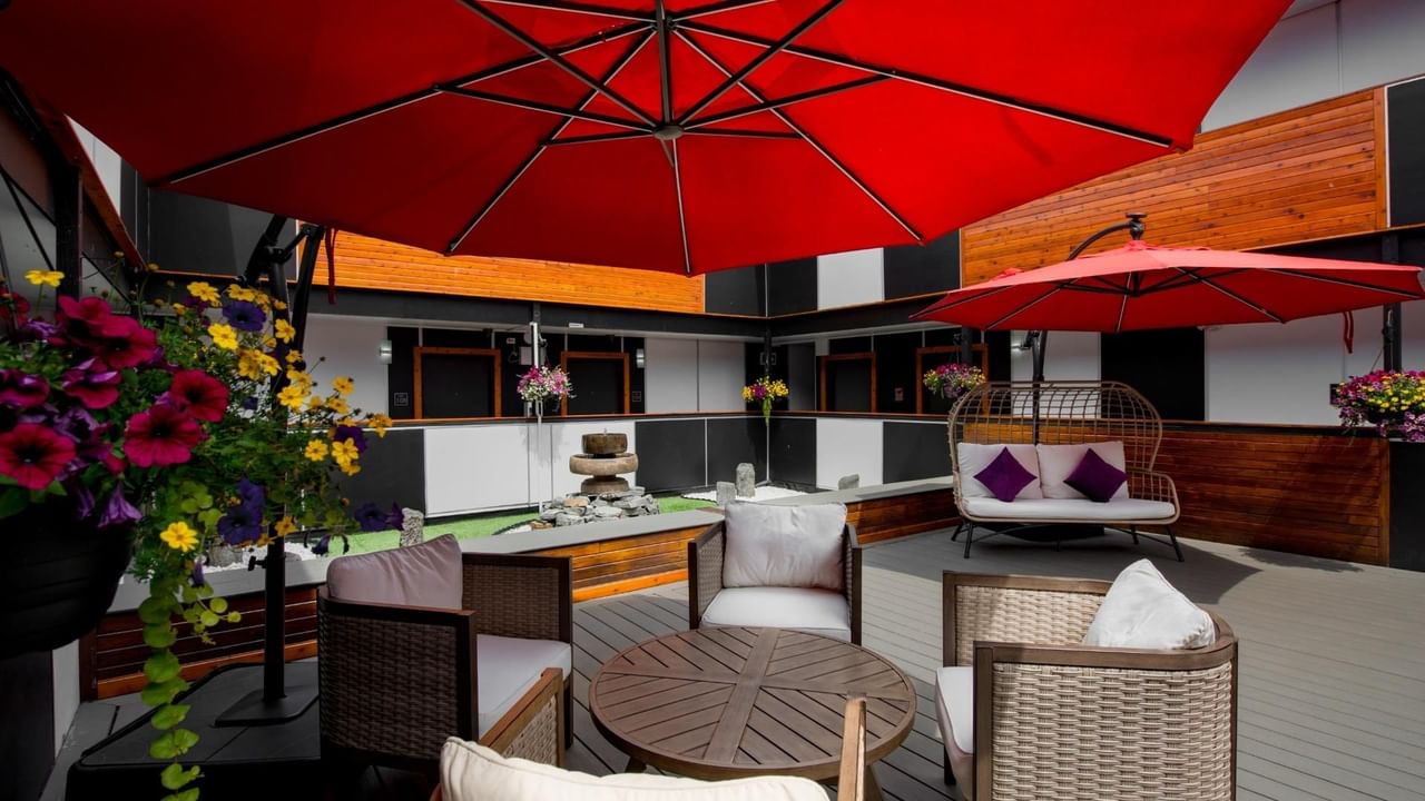 Patio with wicker furniture, red umbrellas, flowers, and a wooden table under a wooden and black facade.