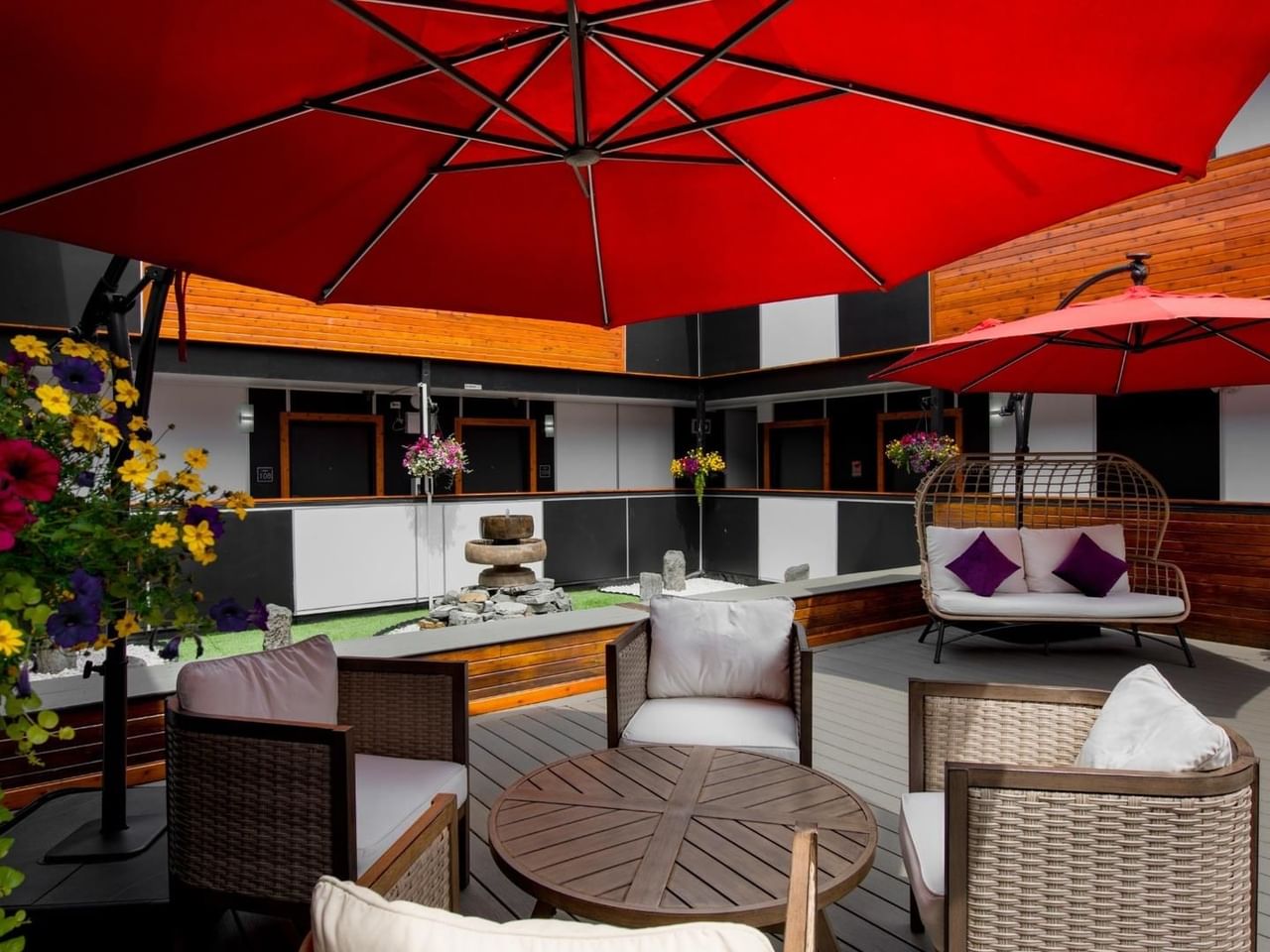 Patio with wicker furniture, red umbrellas, flowers, and a wooden table under a wooden and black facade.