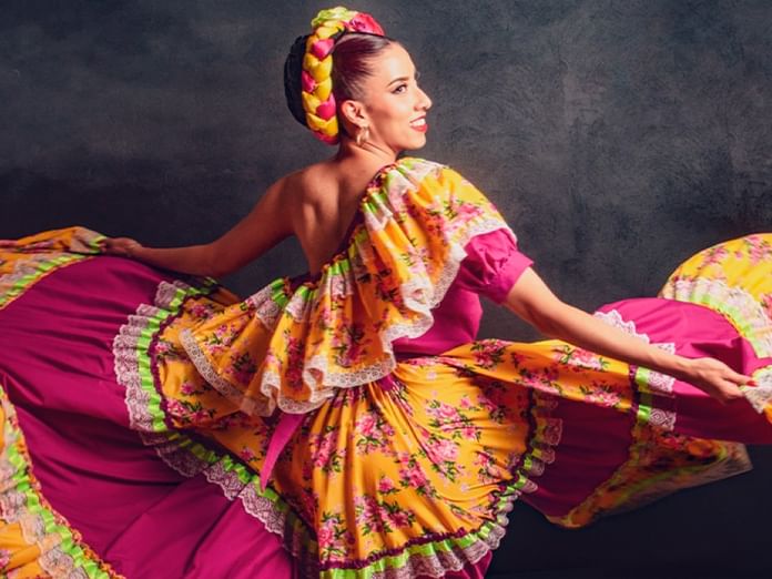 Dancer in vibrant traditional Mexican dress with colorful floral patterns twirling against a dark backdrop.