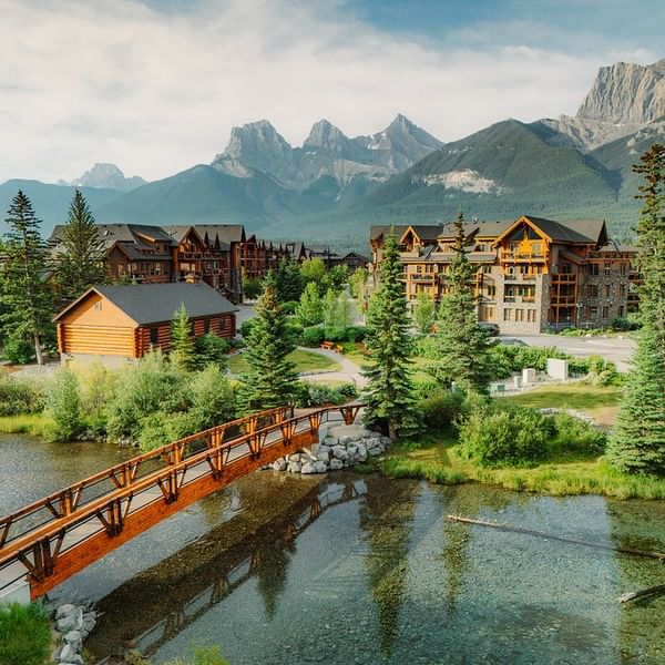 Spring Creek Resorts community with wooden bridge over the creek, surrounded by evergreen trees and dramatic Three Sisters Mountain peaks in the background.