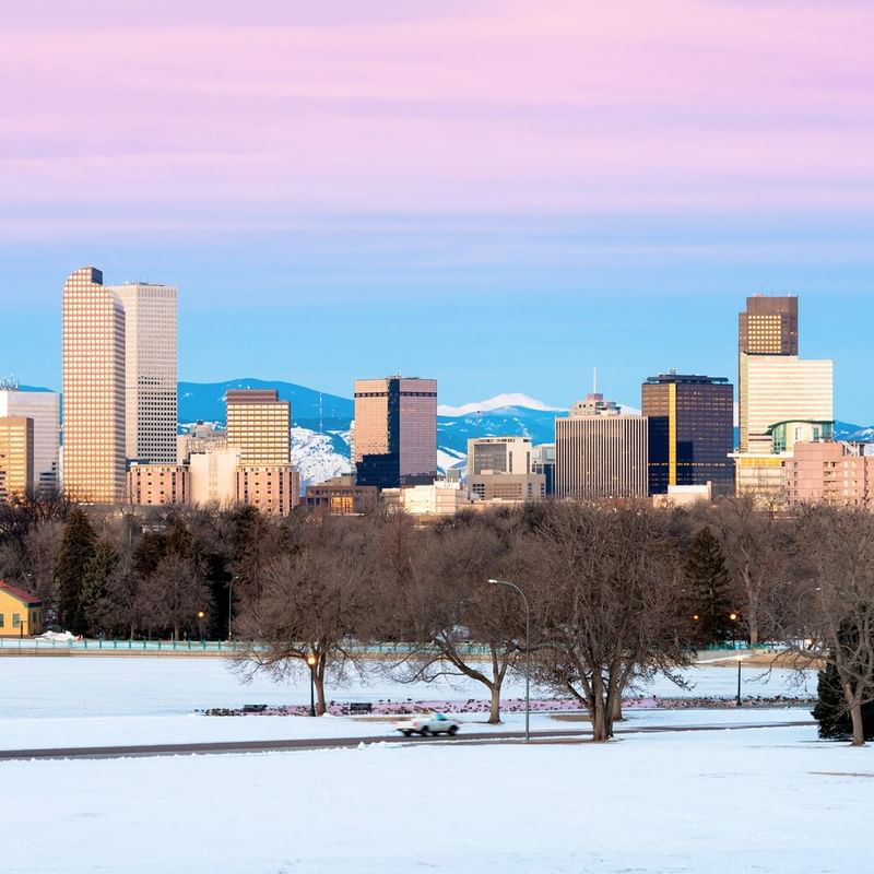 Snowy landscape with a city skyline at dusk, featuring tall buildings and distant mountains.