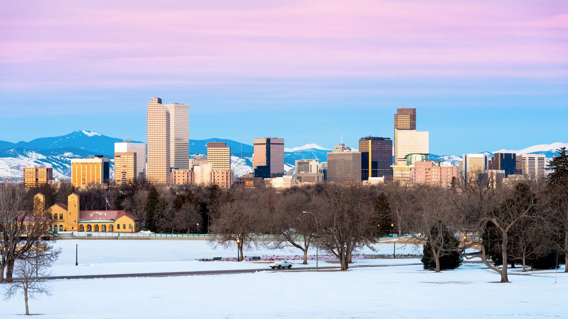 Snowy landscape with a city skyline at dusk, featuring tall buildings and distant mountains.