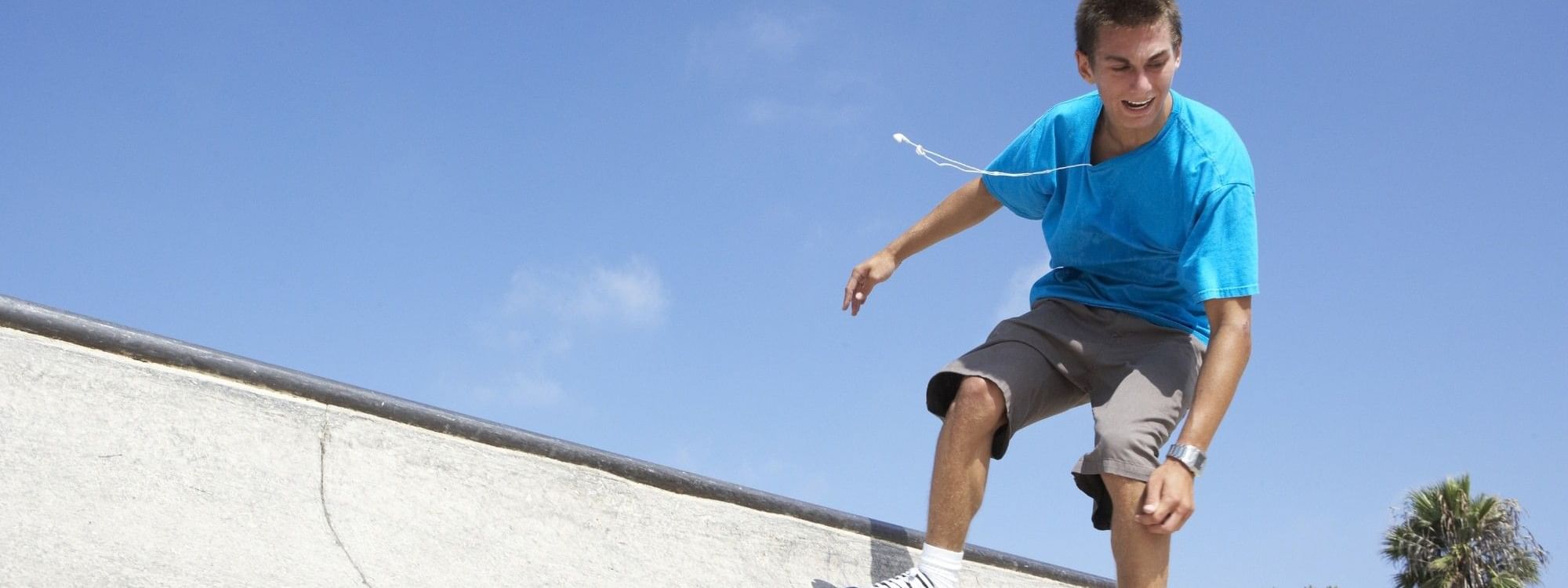 Young skateboarder in blue shirt performing trick on concrete ramp at Haslams Pump Track under clear blue sky.