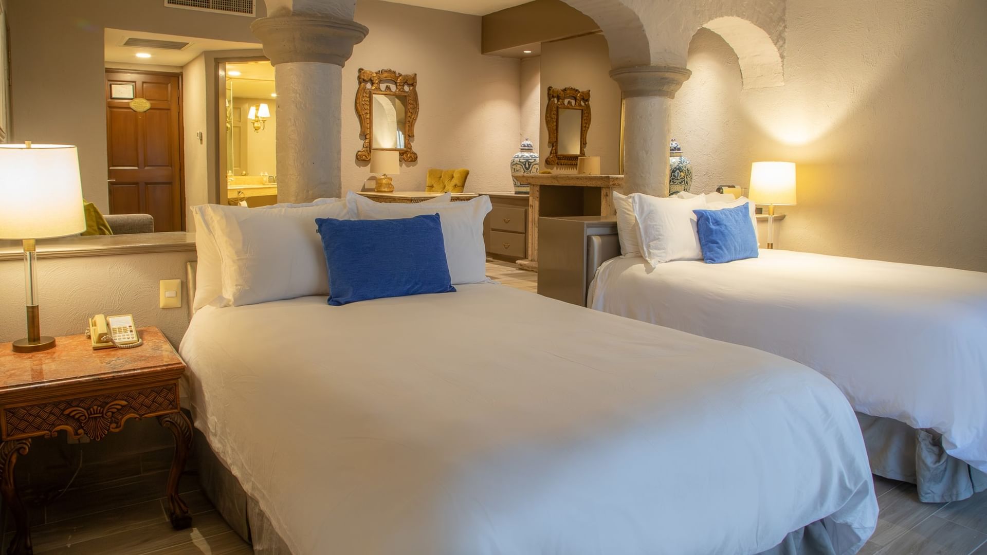 Habitación elegante con dos camas dobles, ropa blanca, cojines azules y arcos coloniales en Quinta Real Guadalajara
