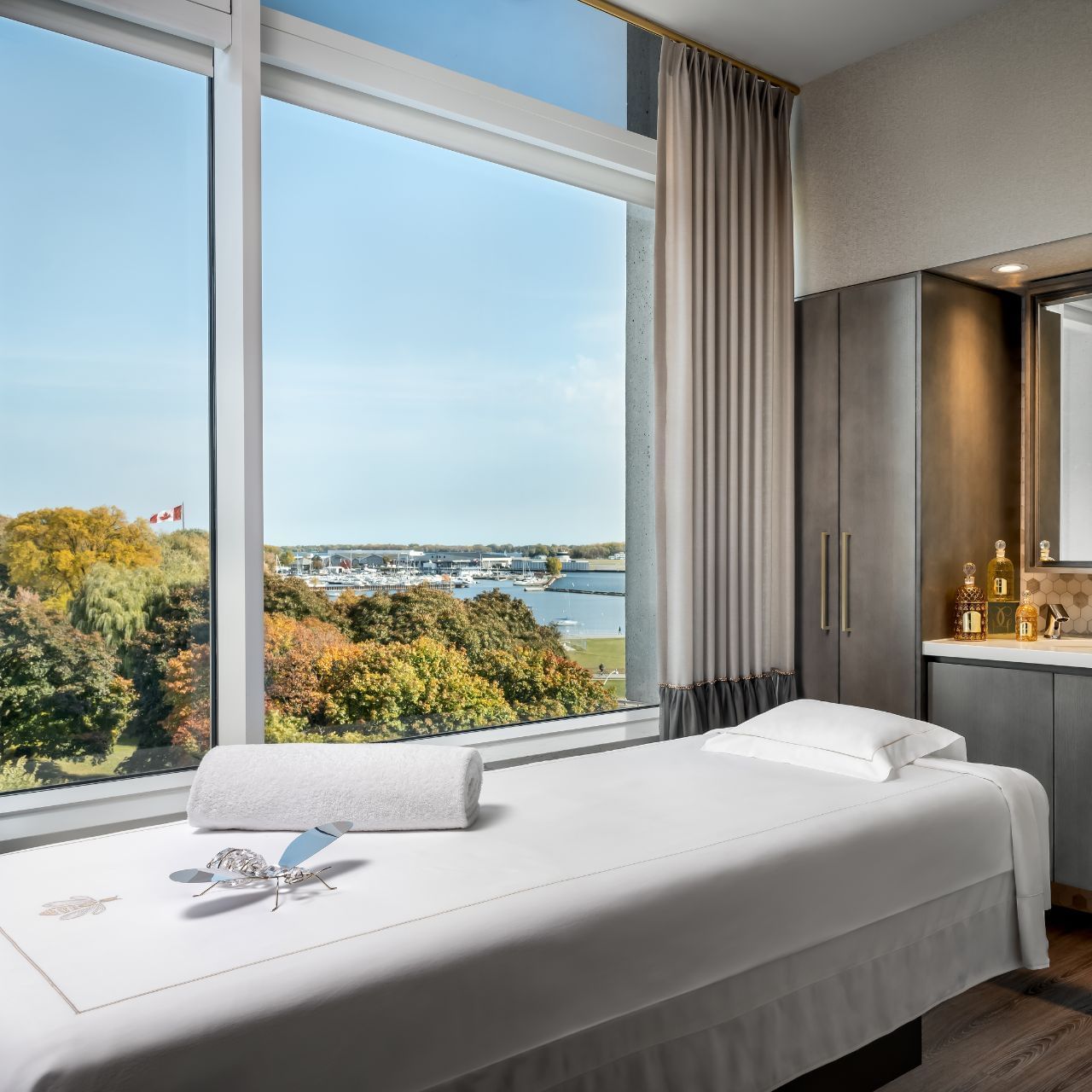 White treatment bed by floor-to-ceiling windows overlooking trees and waterfront in the Guerlain Spa at Hotel X Toronto