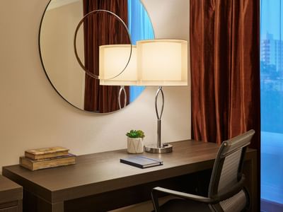 Desk in guest room with chair, lamp, books, and succulent, with a large round mirror and curtains in the background.