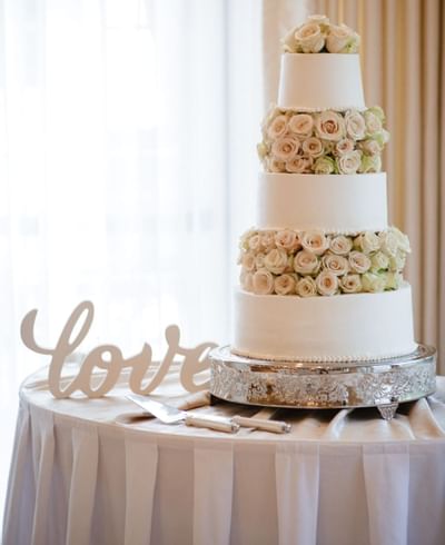 Four tier white wedding cake decorated with fresh pale roses beside a wooden love sign on table at The Simsbury Inn